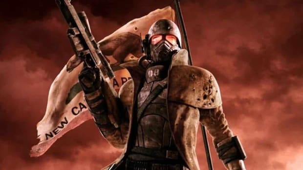 A man in a mask stands with his gun raised, backed by a flag and a red sky in Fallout: New Vegas.