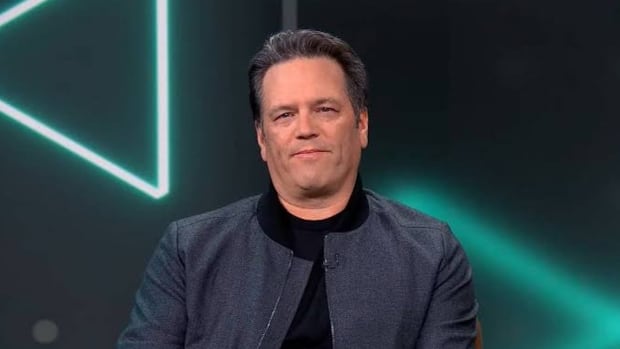 Xbox's Phil Spencer, dressed in a gray coat and black shirt, is sitting on a sofa in front of a green screen decorated with Xbox symbols