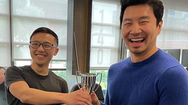 Simu Liu being handed a trophy for winning in a video game.