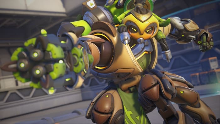 Blizzard plans Overwatch 2 ranked improvements in new seasons: A metal centaur with brown and green plating and a yellow face with green eyes is standing in the middle of a metal room. She's pointing her right arm, which doubles as a laser gun, in front of her