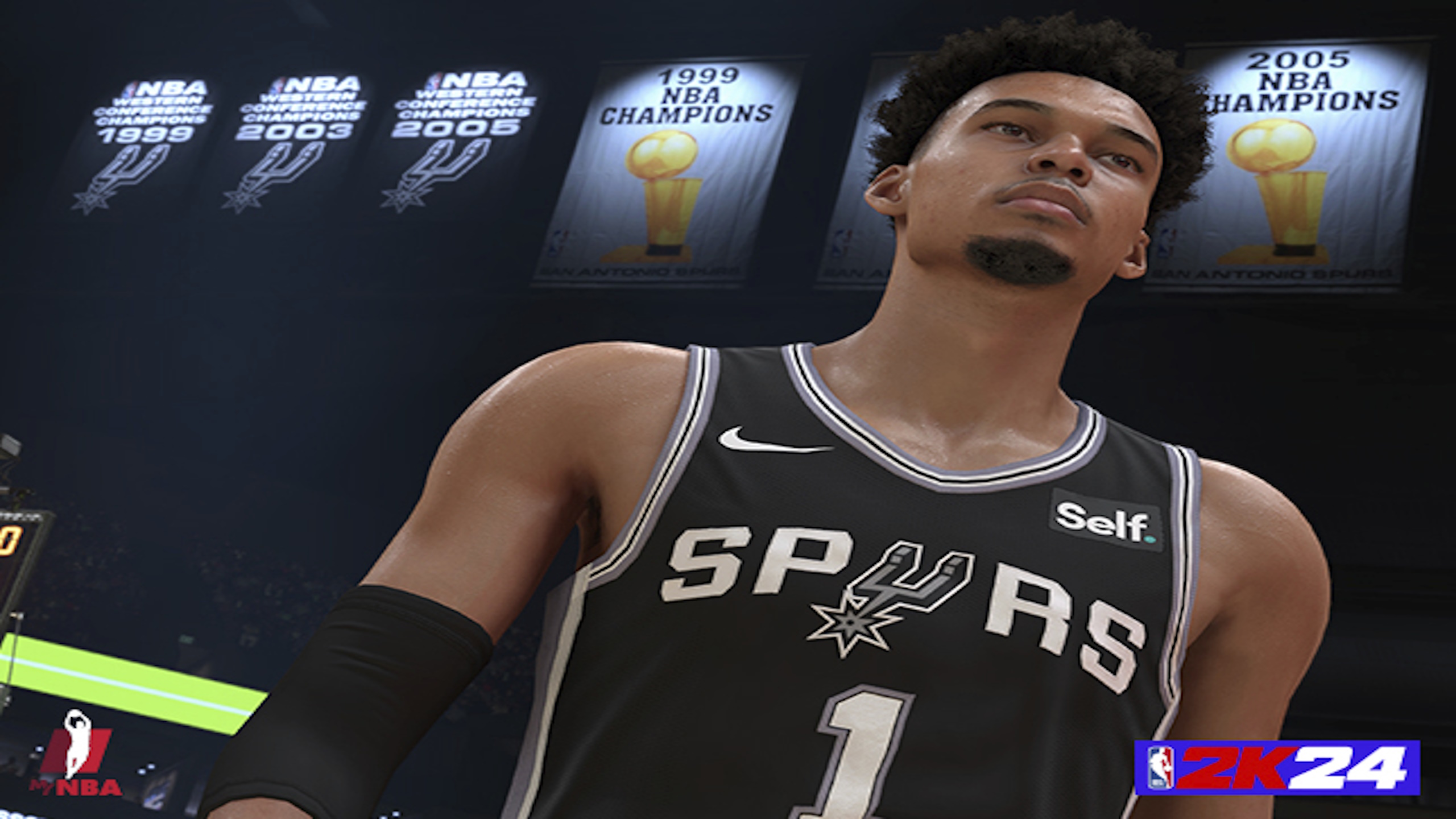 A man is wearing a Spurs #1 jersey while standing on a basketball court with champion pennants all around