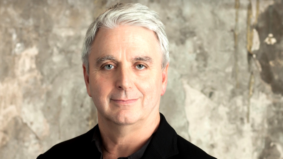 Unity CEO John Riccitiello is depicted - a white man with silvery-white hair, wearing a collared black shirt, is standing in front of a dingy stone wall with a vague smile on his face