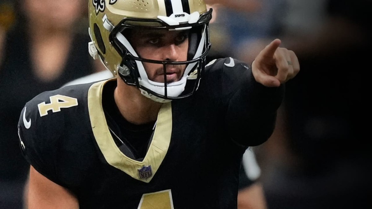 A white man with brown facial hair, wearing a gold NFL helmet and black jersey, is pointing ahead with his left hand.
