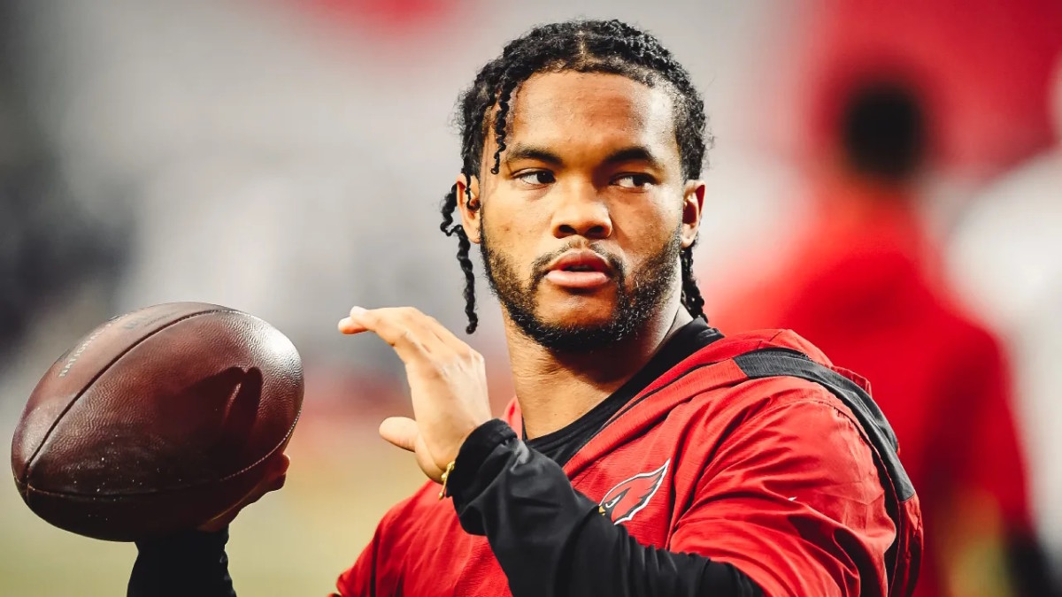 Kyler Murray, a Black man wearing a red Cardinals jersey, is looking to his left, preparing to throw a football with his right hand.
