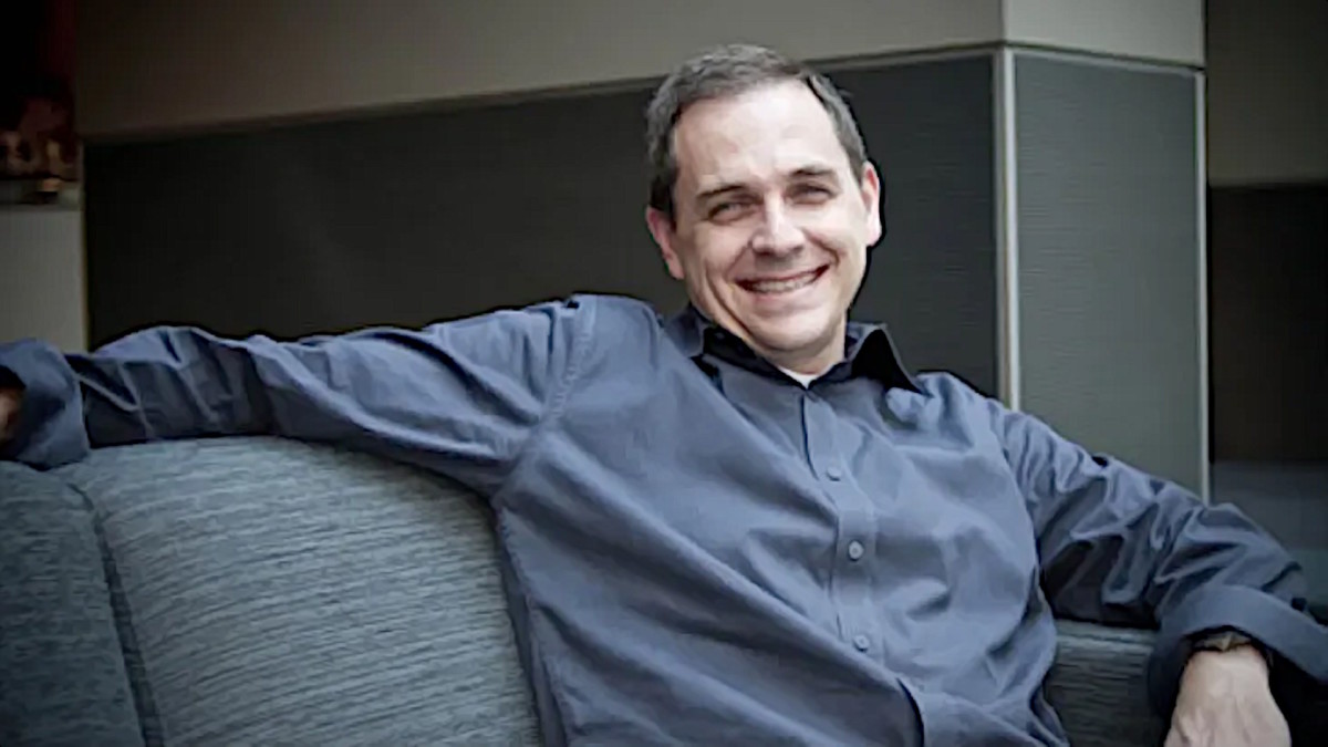 Marc Whitten, a white man with short gray hair, is sitting on a gray sofa, wearing a gray collared shirt, and smiling at the camera