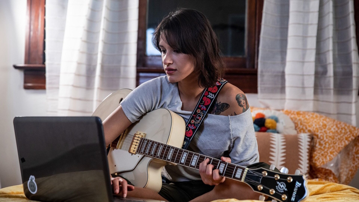 A young woman with shoulder-length dark hair is sitting on a bed with a white guitar on her shoulder and a laptop computer in front of her.