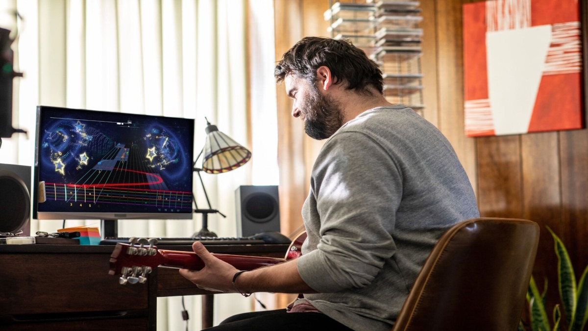 A man in a long-sleeved shirt is sitting at a desk with a guitar in his hand and Rocksmith+ playing on a desktop computer