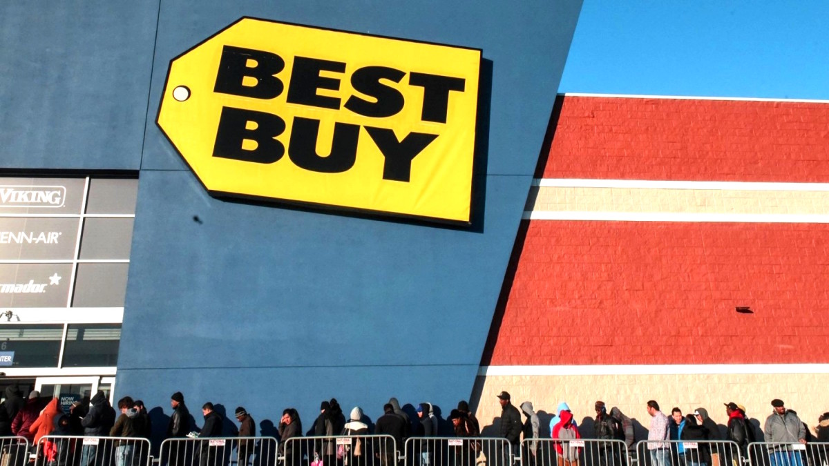 The front of a Best Buy store, with blue paint around the entrance and a yellow price tag sign with "Best Buy" written in large black letters is shown.